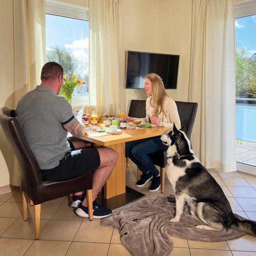 Ferienwohnungen Ostseeresidenz Schönberger Strand, Küchenbereich mit Esstisch und zwei Stühlen, Pärchen sitzt sich am Tisch gegenüber und frühstückt, an der Wand TV-Gerät, vor dem Tisch sitz Hund auf Decke, rechts Zugang zum Balkon