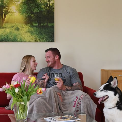 Ferienwohnung Ostseeresidenz Schönberger Strand, Wohnzimmer, lächelndes Pärchen sitzt auf dem roten Sofa mit einer Decke über den Beinen, jeder hat ein Glas Orangensaft in der Hand, Vordergrund Glastisch mit Vase und Tulpen, daneben sitzender Hund