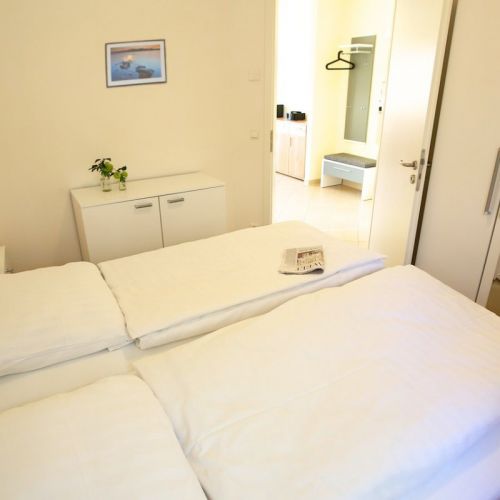 Ferienwohnungen Ostseeresidenz Schönberger Strand, Schlafzimmer mit Doppelbett, an der Wand Sideboard, darüber Bild, Blcik aus dem Schlafzimmer in den Eingangsbereich mit Gaderobe und Sitzmöglichkeit