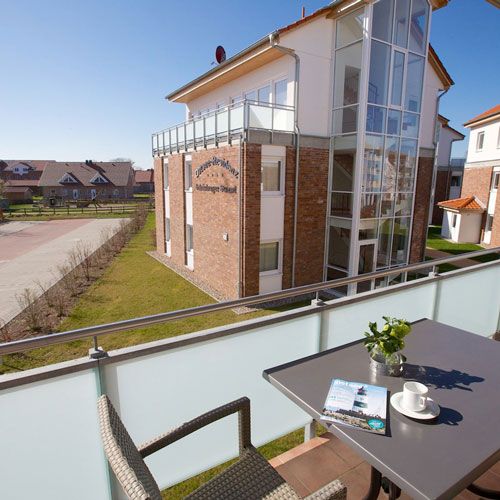 Ferienwohnungen Ostseeresidenz Schönberger Strand, Blick vom Balkon auf ein weiteres Gebäude, Rattanstuhl und Tisch, auf dem Tisch Kaffeetasse mit Unterteller, Zeitschrift, Vase mit Blumen