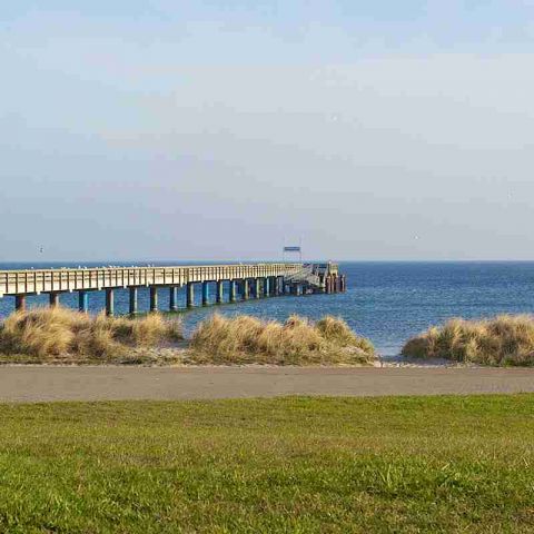 Ostseeresidenz Schönberger Strand, im Vordergrund Promenade mit Grünstreifen, im Hintergrund Seebrücke sowie Düne mit Gräsern und Ostsee