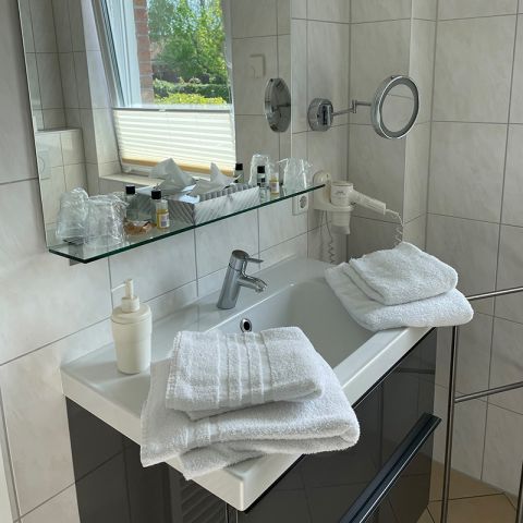 Ferienwohnungen Ostseeresidenz Schönberger Strand, Badezimmer, Waschblock mit Waschbeckern und Handtüchern, Kosmetikspiegel, Föhn, Spiegel, auf der Spiegelablage Kosmetikartikel, im Spiegel spiegelt sich das angekippte Fenster mit Plissee