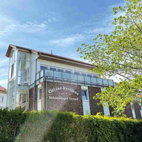 Ostseeresidenz Schönberger Strand, Ansicht eines Gebäudes mit der Beschriftung Ostseeresidenz Schönberger Strand an der Hauswand, im Vordergrund grüne hecke und hereinragender Baum