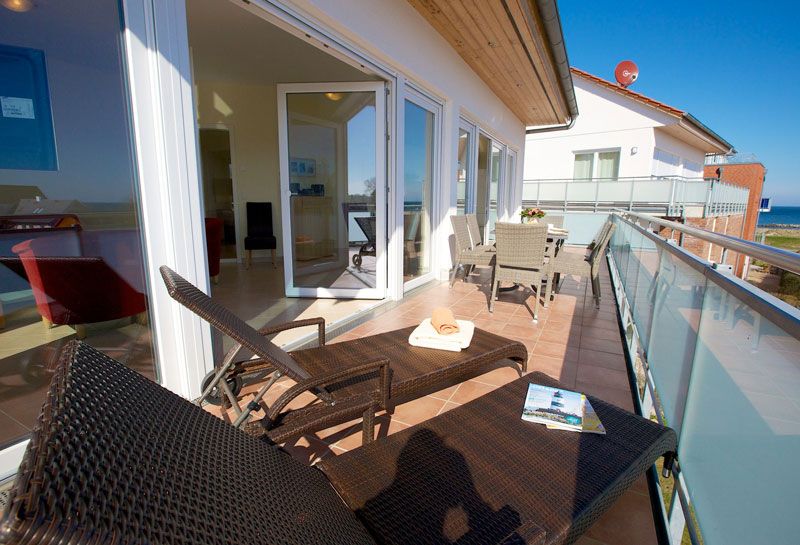 Penthouse Ostseeresidenz Schönberger Strand, Balkon mit zwei Rattanliegen sowie sechs Rattanstühle und Tisch, Tisch ist mit Frühstück eingedeckt, geöffnete Balkontüren, Blick in den Wohn- und Essbereich