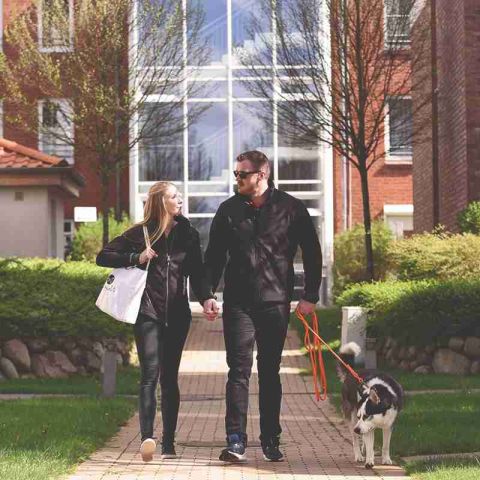 Ostseeresidenz Schönberger Strand, Pärchen mit Hund geht zwischen den Gebäuden der Ostseeresidenz Schönberger Strand spazieren, Pärchen schaut sich an, Hund läuftan der Leine, Frau trägt weiße Tasche