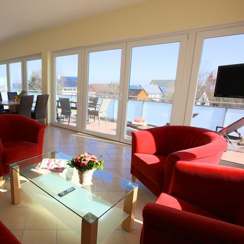 Penthouses Ostseeresidenz Schönberger Strand, Wohn- und Essbereich, Sitzecke mit drei roten Sesseln, zwischen Sesseln steht ein Glastisch, auf Tisch Blumenstrauß, Zeitschriften, Fernbedienung, Essbereich Tisch und 4 Stühlen, Blick auf Balkon