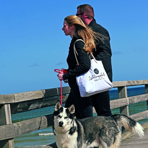 Umgebung Ostseeresidenz Schönberger Strand, Strand, Sand, Seebrücke, Ostsee, Pärchen am Geländer der Seebrücke mit Blick auf die Ostsee, Hund an der Leine, Frau trägt weiße Tasche mit Aufdruck Anders am Meer Lieblingsplatz