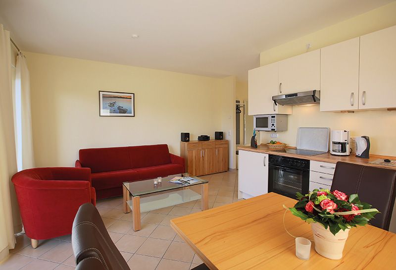 Ferienwohnung Ostseeresidenz Schönberger Strand, Wohnzimmer mit Küche und Essbereich, an der Wand rotes Sofa und roter Sessel mit Glastisch, über dem Sofa Bild, neben dem Sofa Sideboard mit Musikanlage, rechts Küchenzeile mit Küchengeräten
