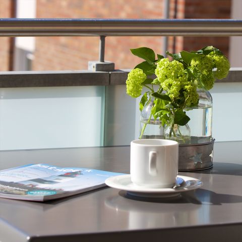 Ferienwohnung Ostseeresidenz Schönberger Strand, Ausschnitt Balkon, Nahaufnahme Tisch mit Zeitschrift, Kaffeetasse auf Unterteller und Vase mit Blumen, m Hintergrund Balkongeländer